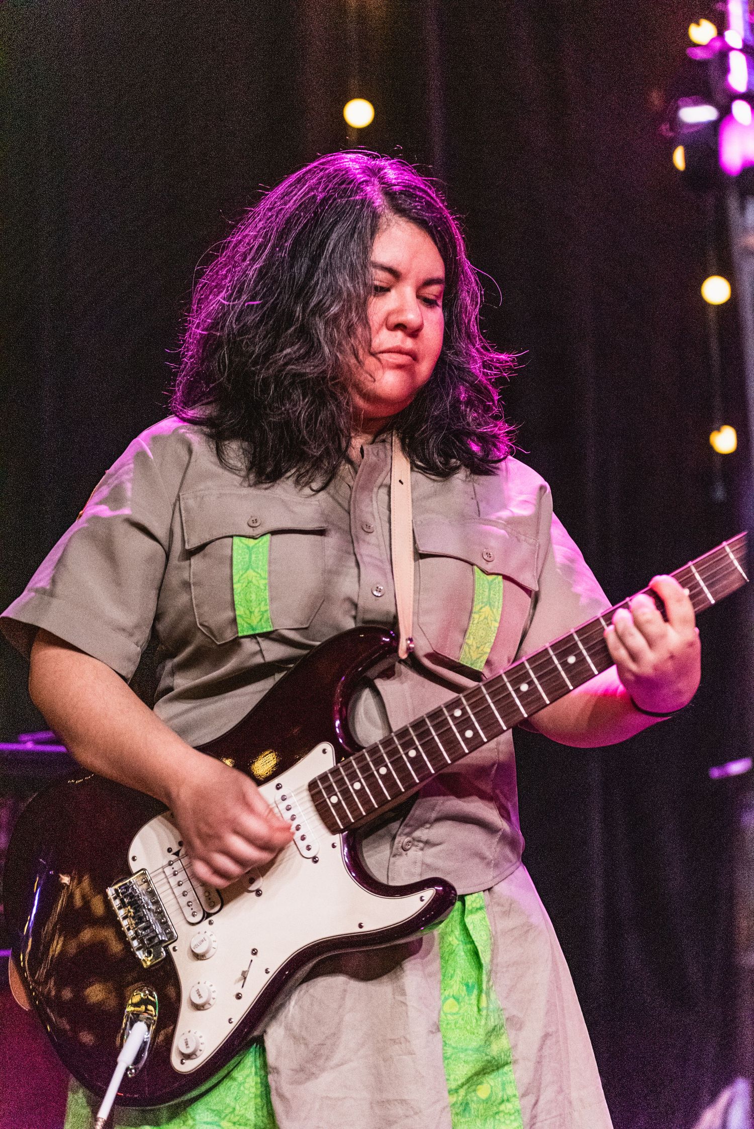 Musician plays white Stratocaster guitar while wearing khaki uniform with green accents under stage lights.