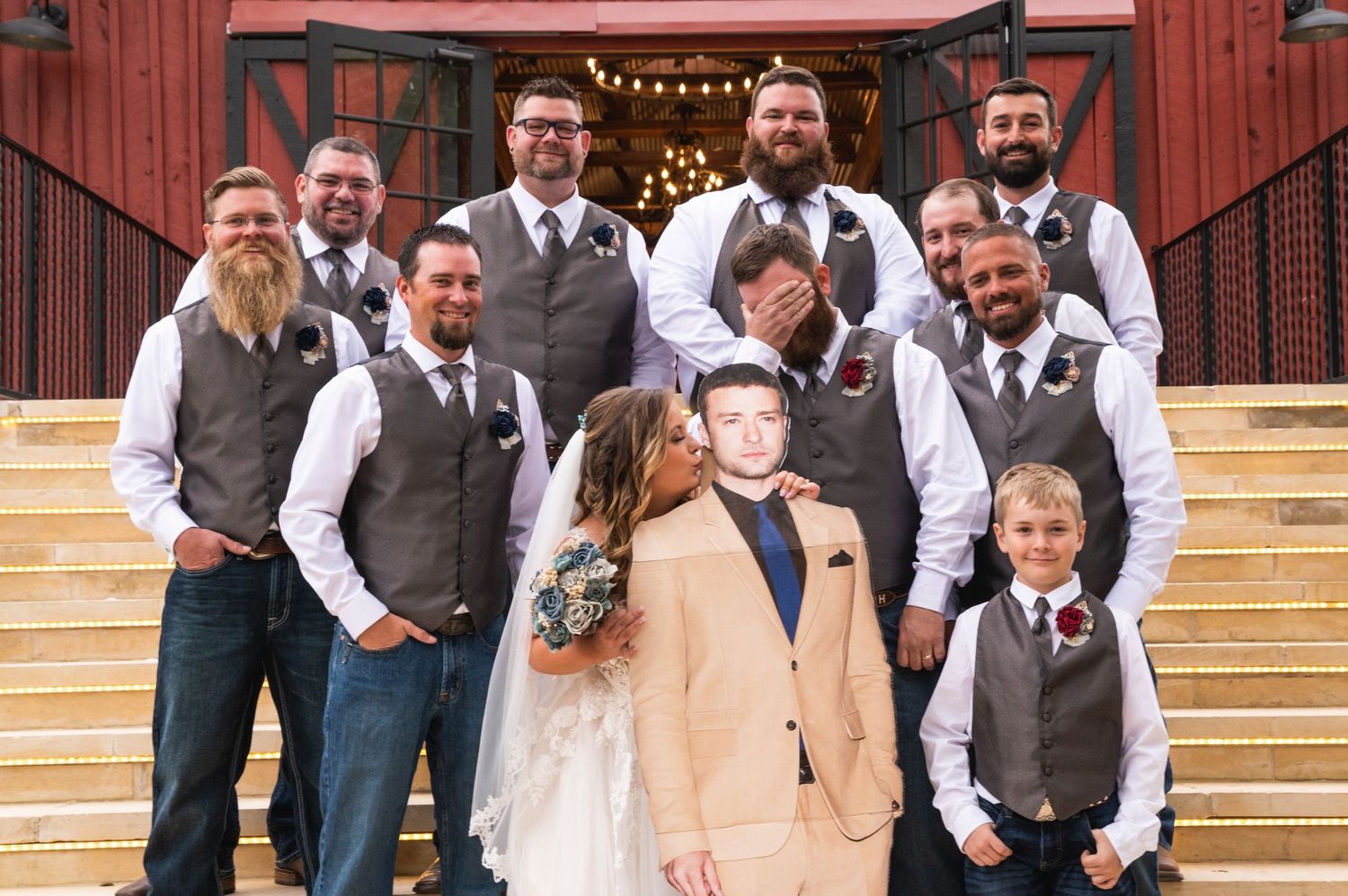 Fun group photo of wedding party posing on stairs in front of red barn venue.