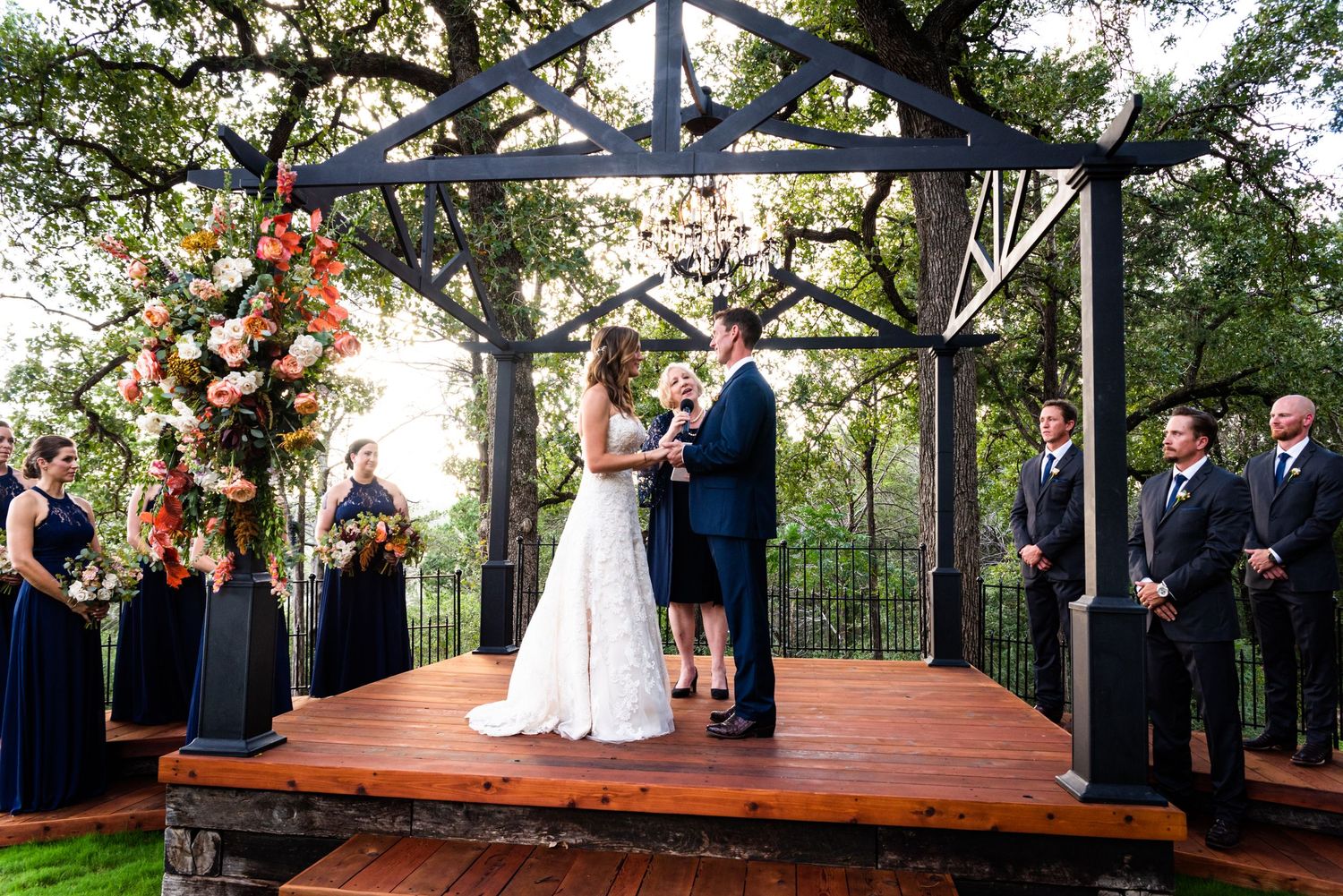 Wedding ceremony on wooden deck with floral arch and wedding party in navy attire.