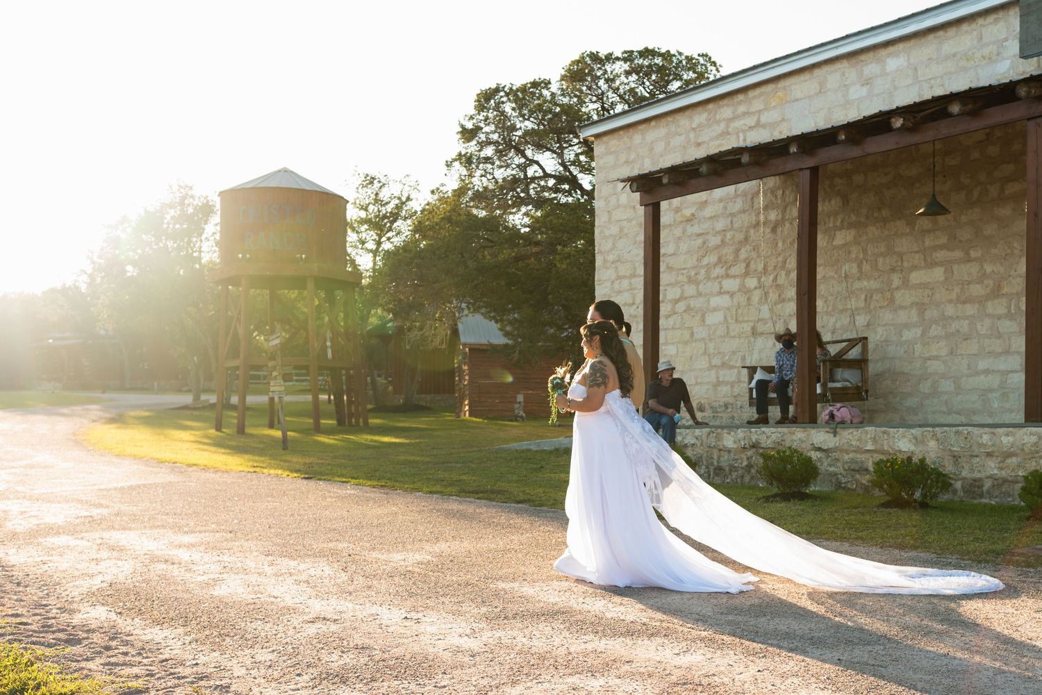 Bride in flowing white gown poses at sunset outside rustic stone building.