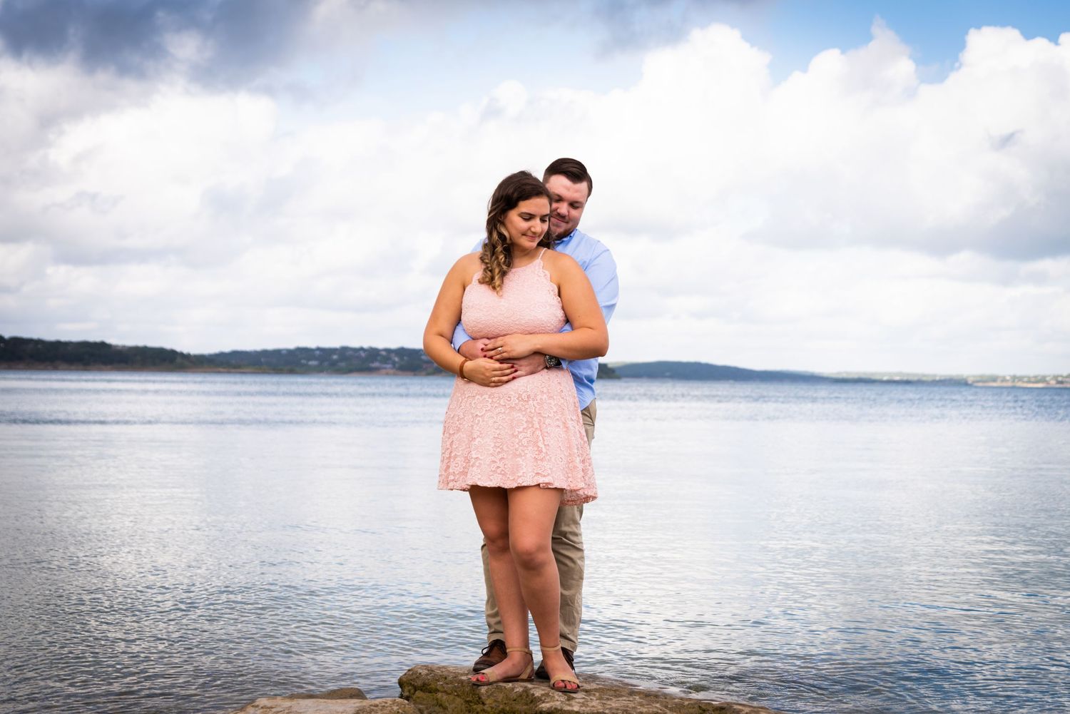 A couple embraces while standing on rocks with a scenic water view behind them.
