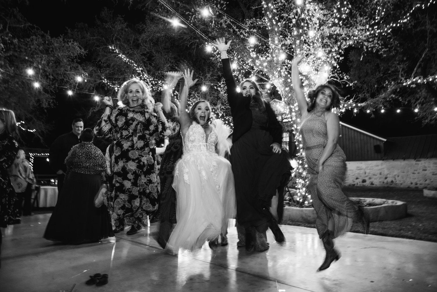 A black and white photo of wedding guests dancing under illuminated trees.