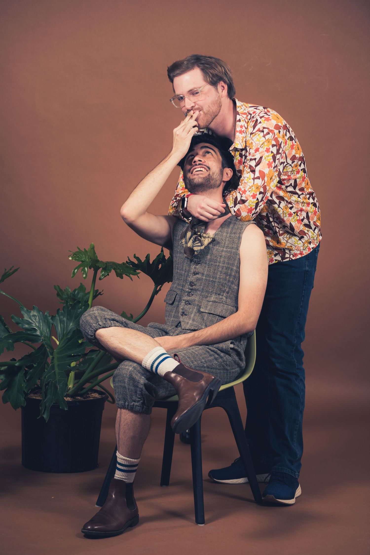 Two people pose playfully in a studio setting with potted plants and a brown backdrop.