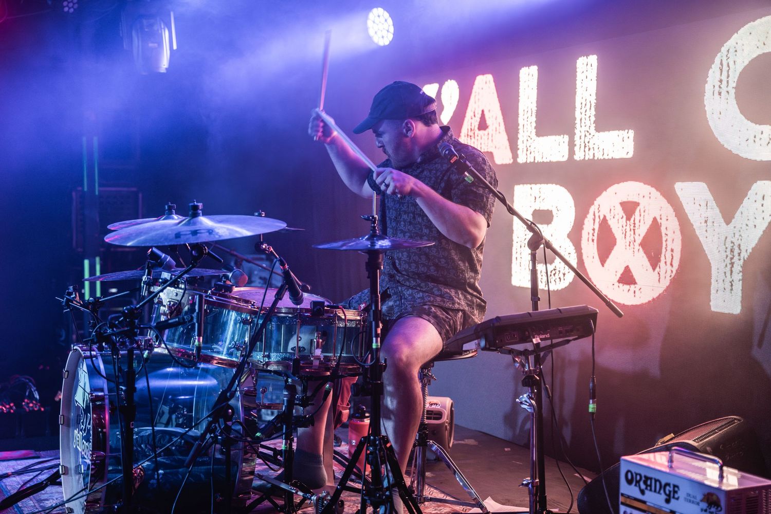 Drummer performs energetically on stage with illuminated Fall Out Boy logo and purple lighting in the background.