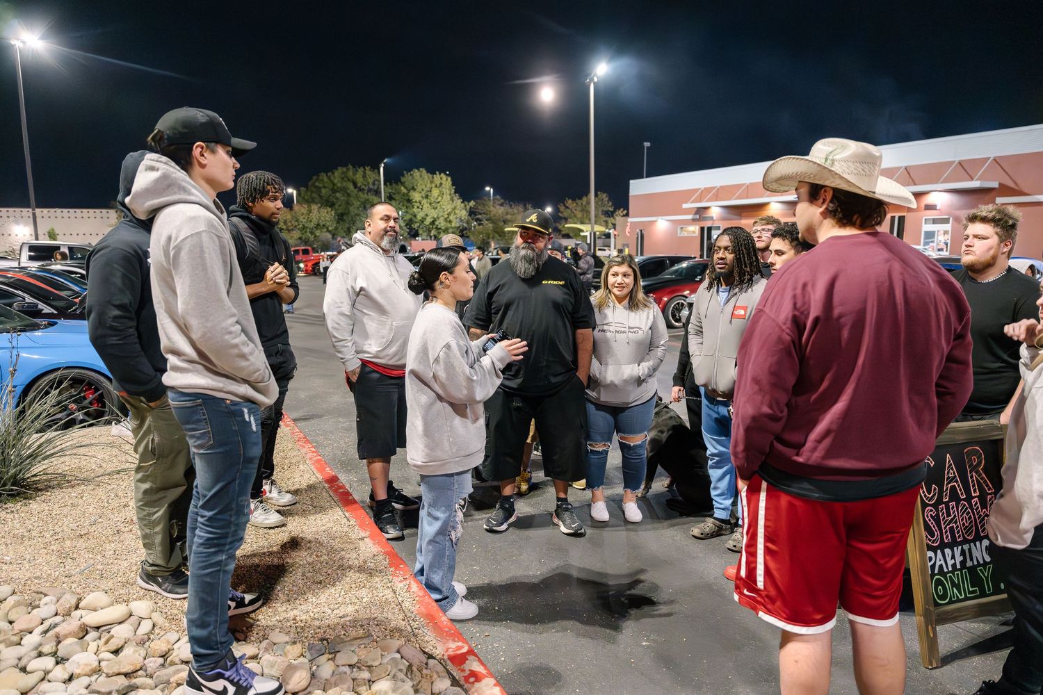 Group of car enthusiasts gathered in parking lot at night car meet.