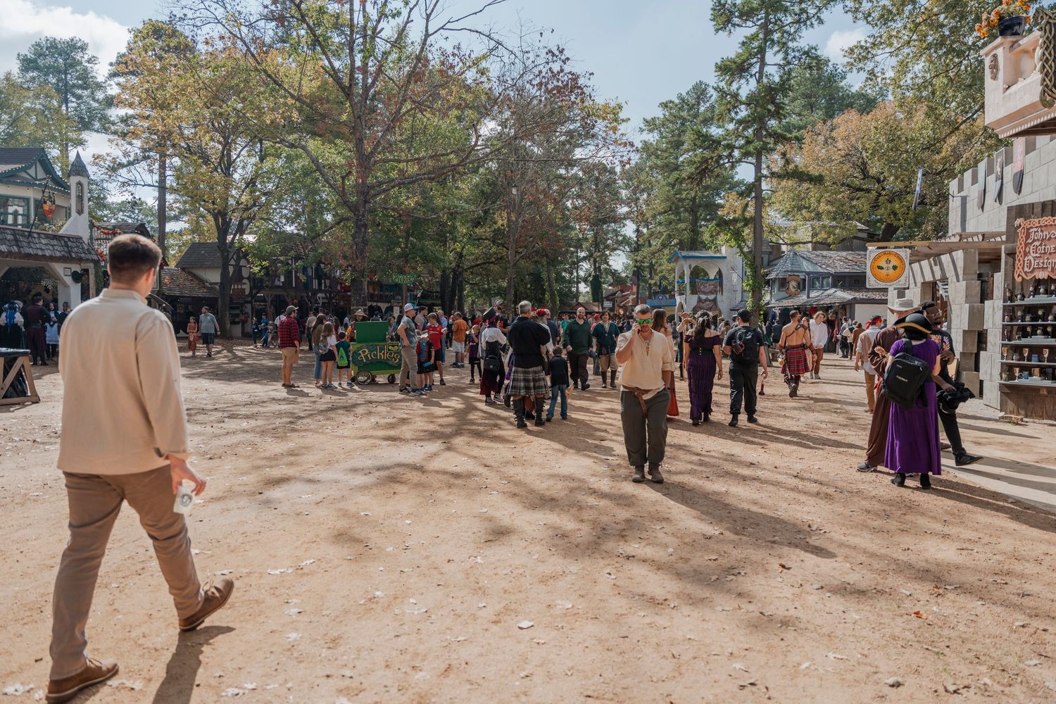 Dirt path lined with vendor stalls and festival attendees in medieval costumes at an outdoor renaissance fair.