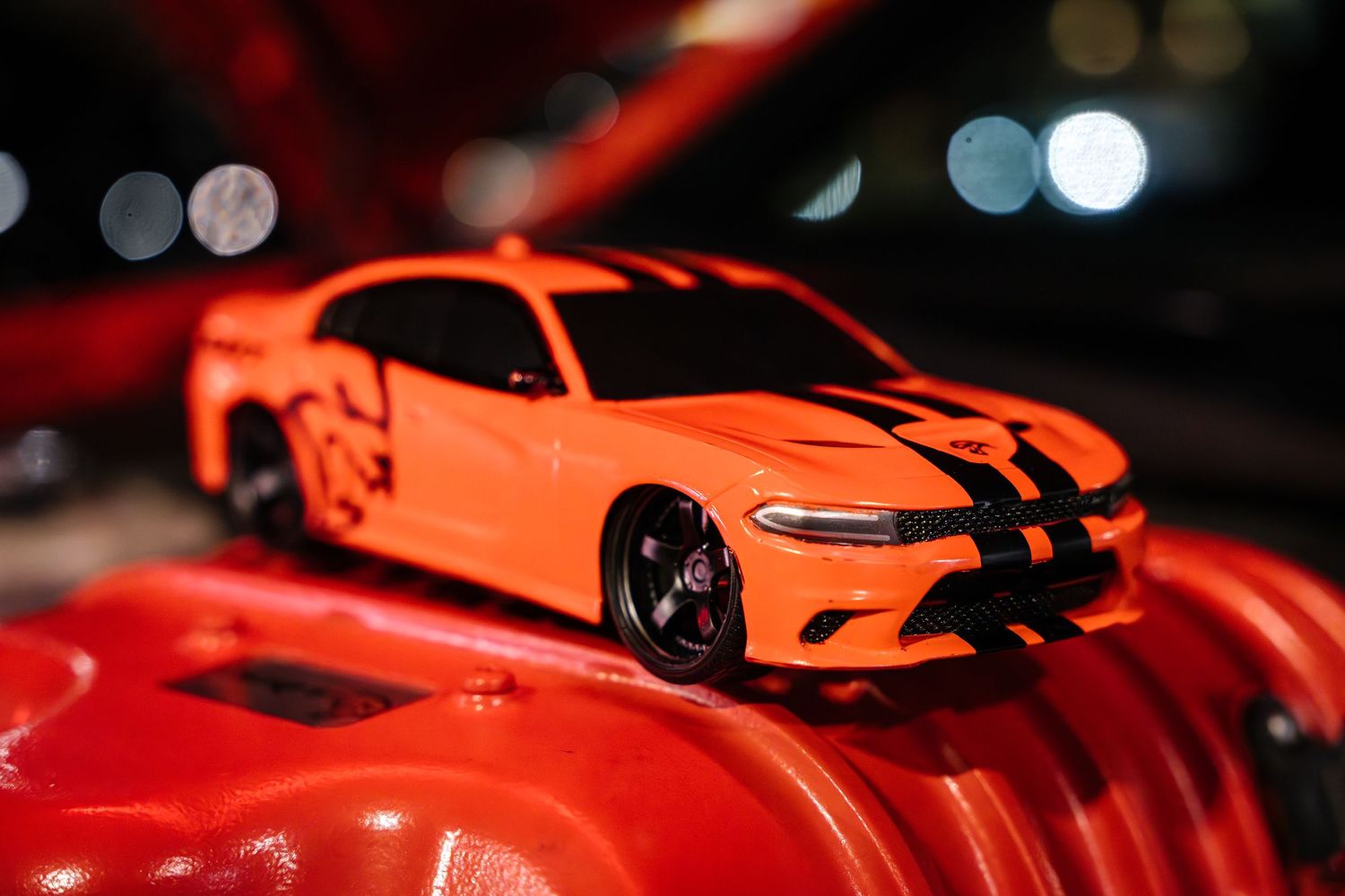 Orange Dodge Charger model car with black racing stripes displayed on red surface.