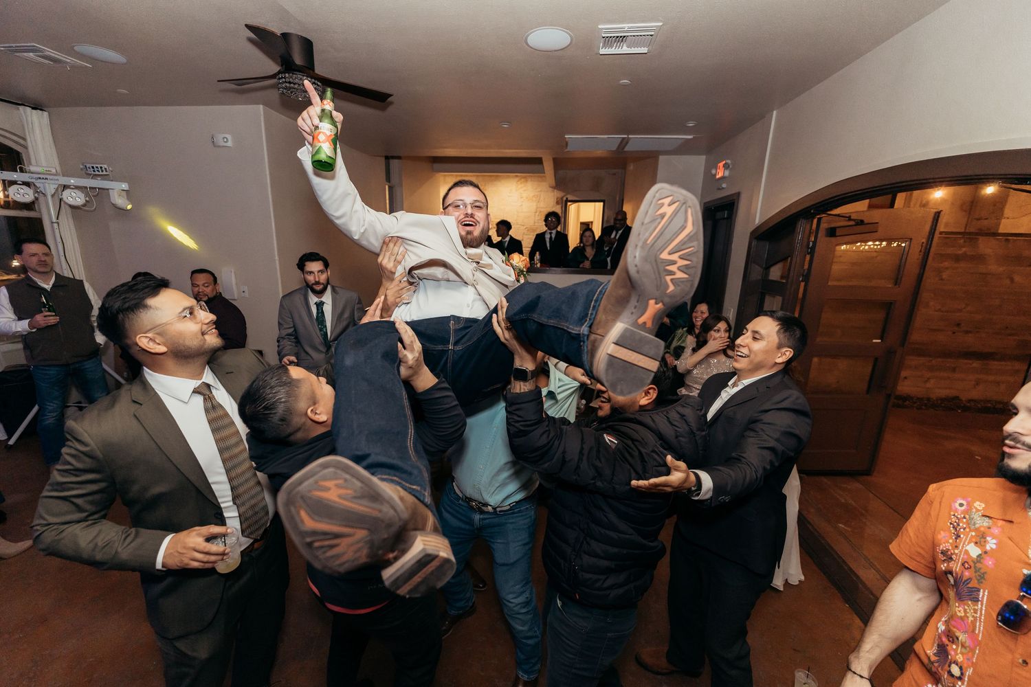 Groomsmen lifting and celebrating with someone at an indoor wedding reception.