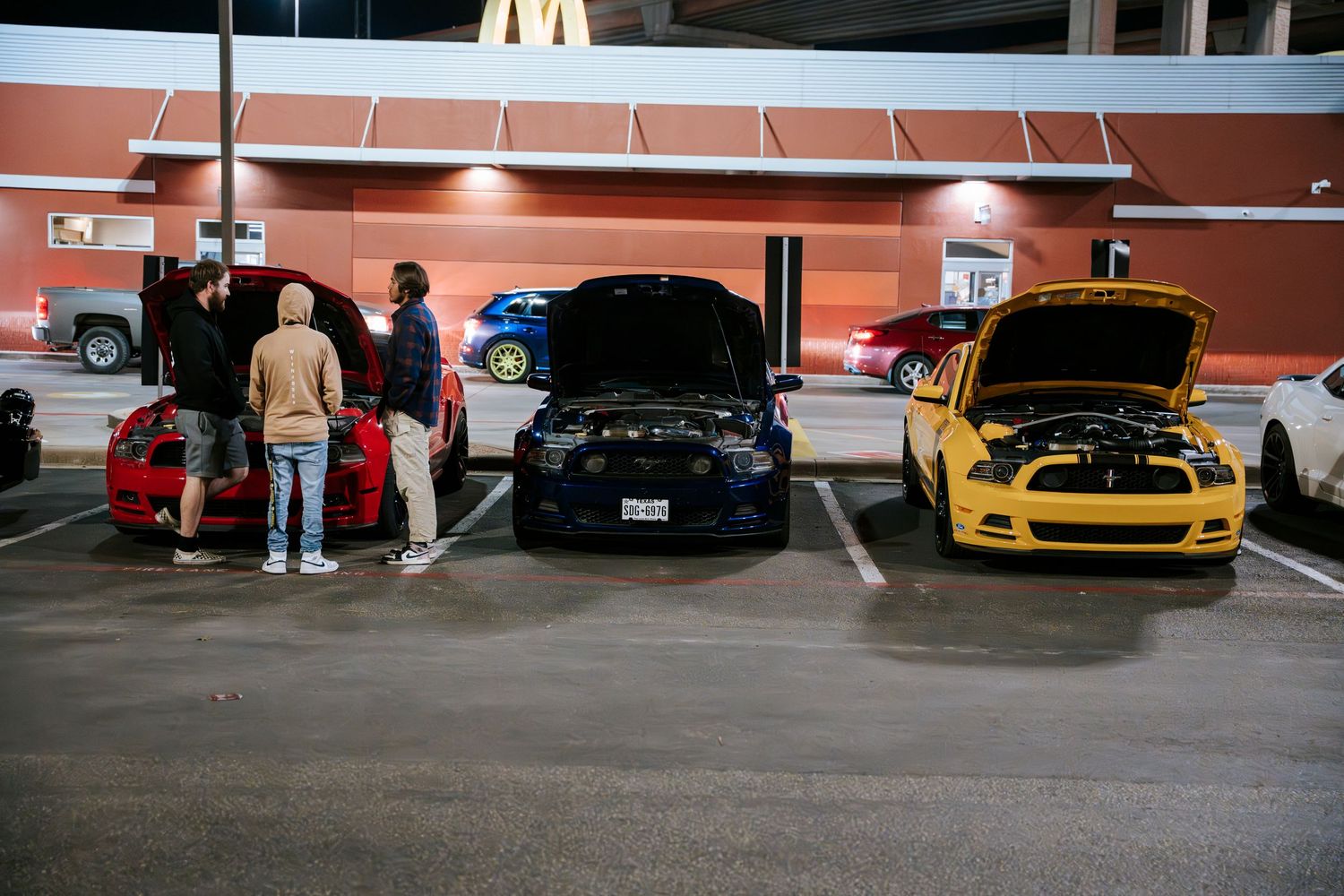 Performance cars lined up at a nighttime car meet with hoods open for display.