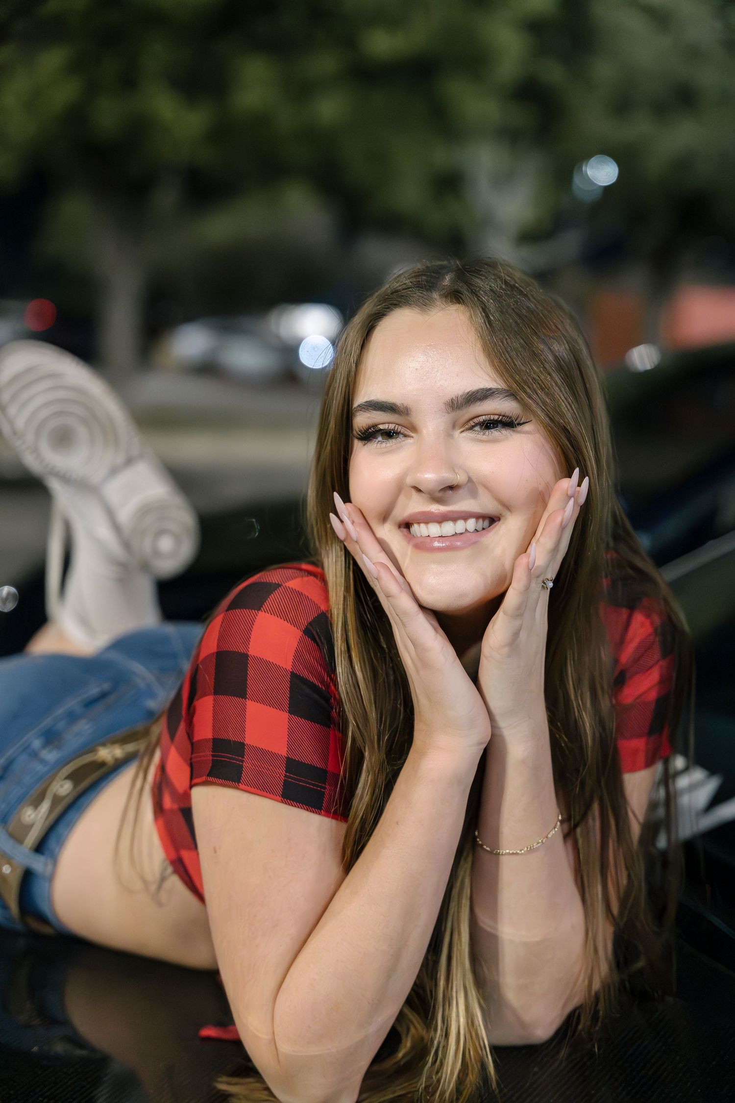 Someone in red buffalo plaid poses on car hood at night.