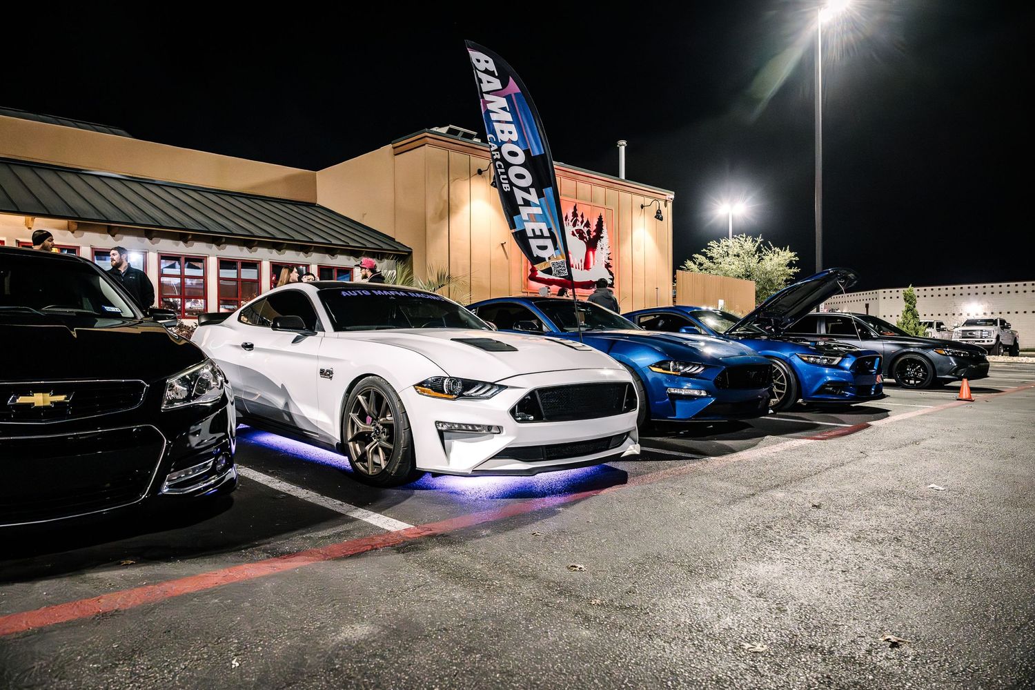 Modified sports cars including Mustangs and other performance vehicles on display in a nighttime parking lot.