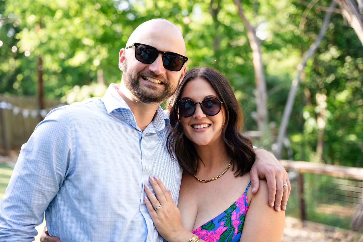 A couple wearing sunglasses smiles outdoors on a sunny day with green foliage in the background.