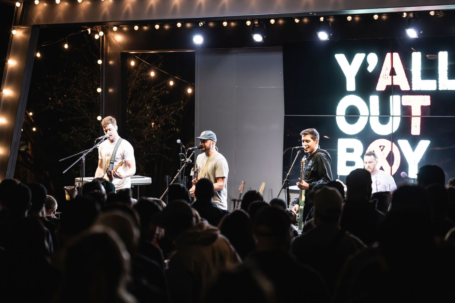 Band performs on stage under bright lights with 'Y'ALL OUT BOY' text display in background.