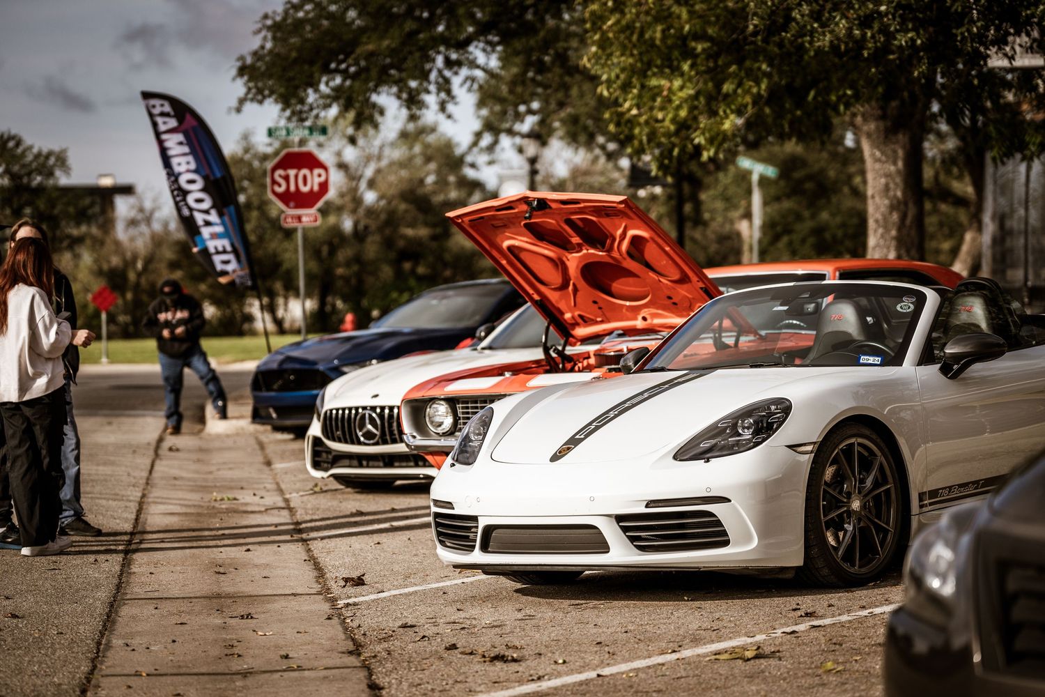 A white Porsche and Mercedes-Benz displayed at a car show with a promotional flag banner visible in the background.