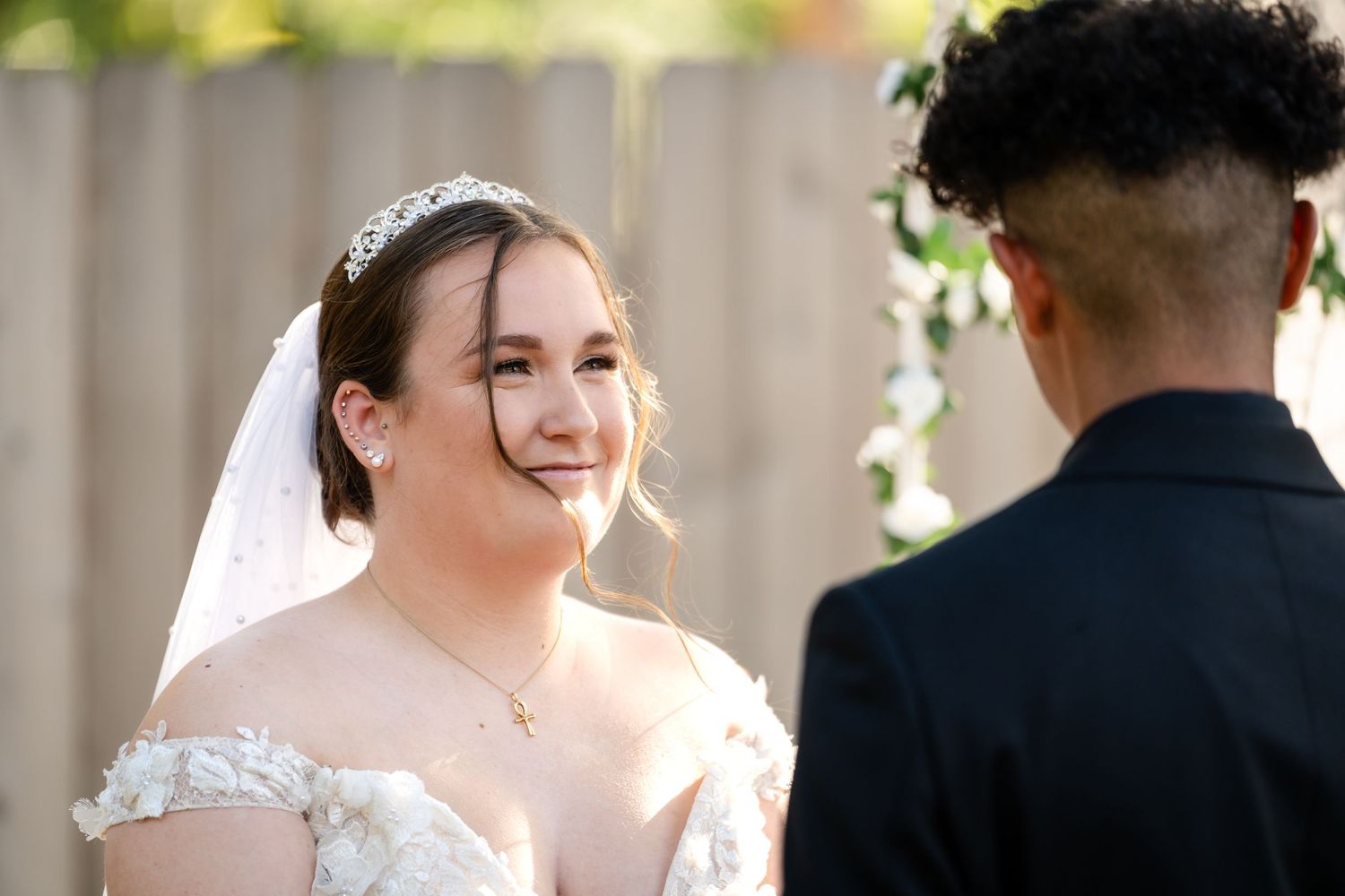A bride in a white lace dress smiles joyfully during her outdoor wedding ceremony.