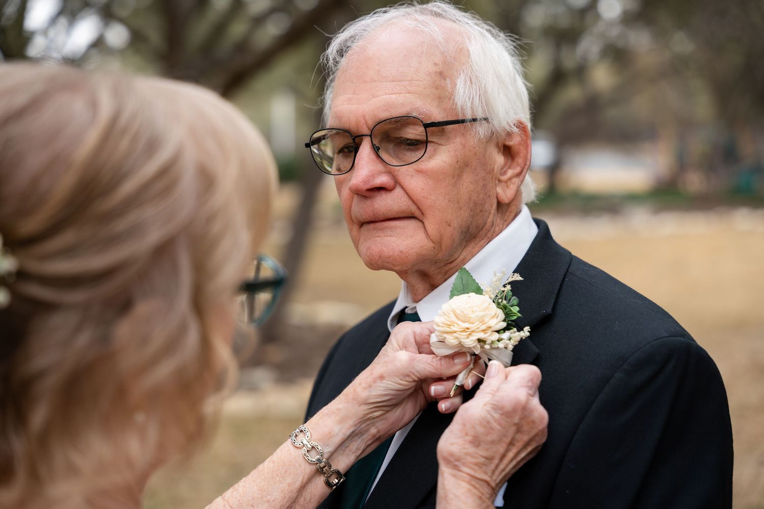 Someone adjusts a pink and white floral boutonniere on a man wearing glasses and a black suit at an outdoor event.