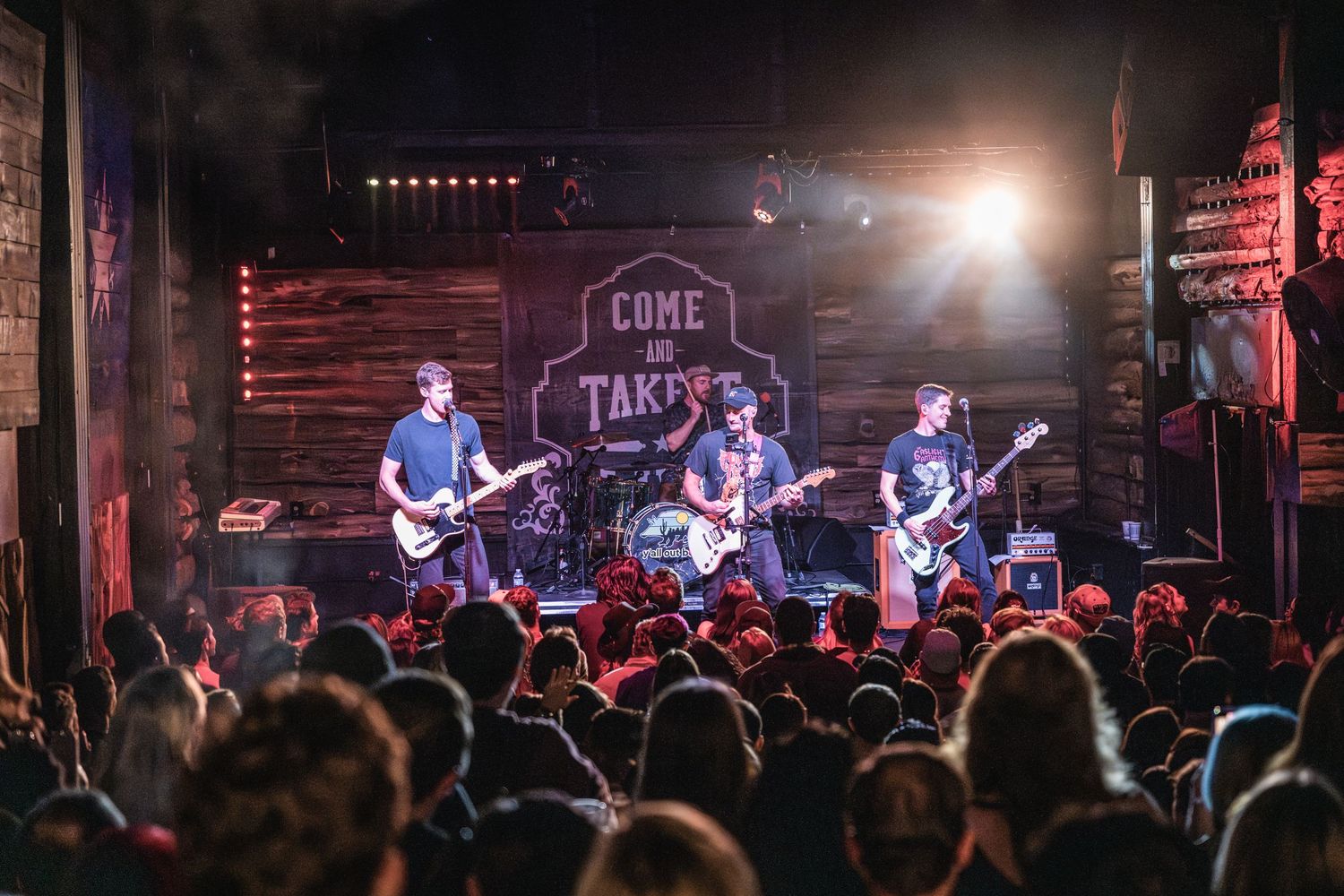 Band performing at packed venue with 'Come Take It' sign visible in background.