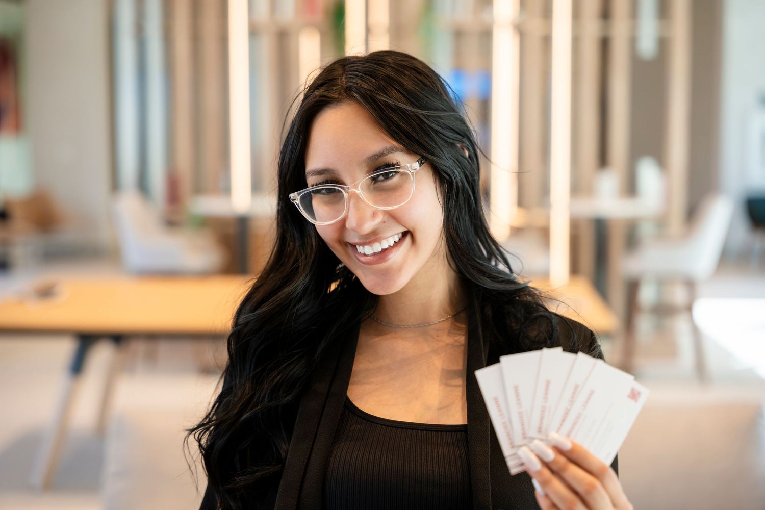 Someone with long dark hair and glasses smiles while holding up playing cards in an office setting.