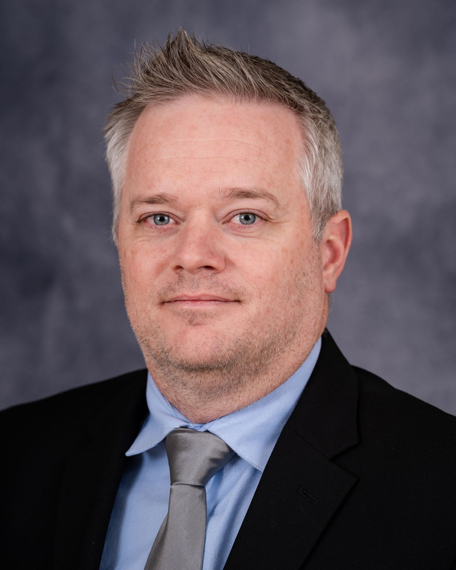 Professional headshot of a person in suit and tie with gray mottled background.