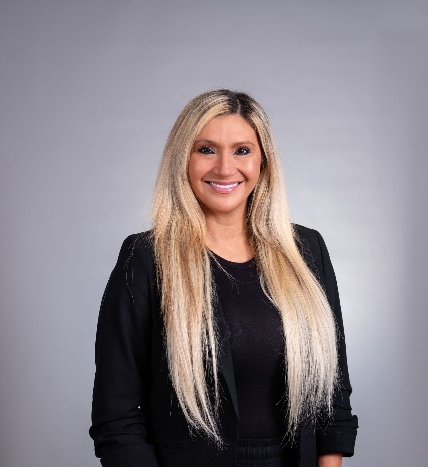Professional headshot of person in black top with long blonde hair smiling against grey background.