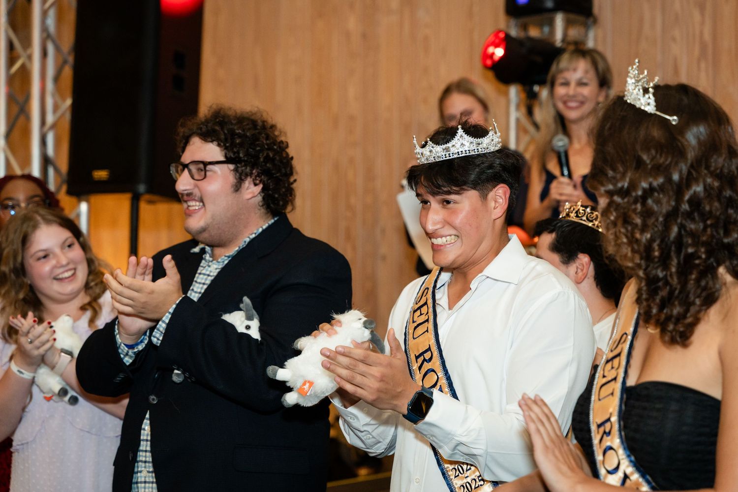 Participants celebrate during a festive indoor event wearing formal attire and decorative sashes.