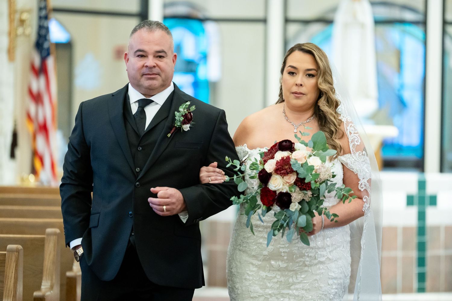 Wedding procession walking down church aisle with bridal bouquet featuring deep red flowers.