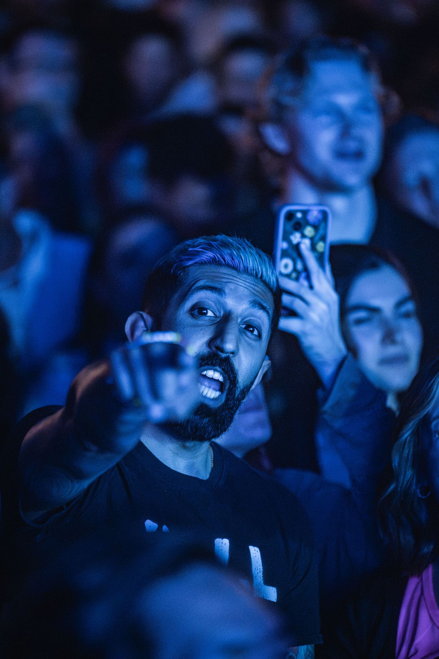 Concert audience members in blue lighting watch a performance while holding up mobile phones to record the event.