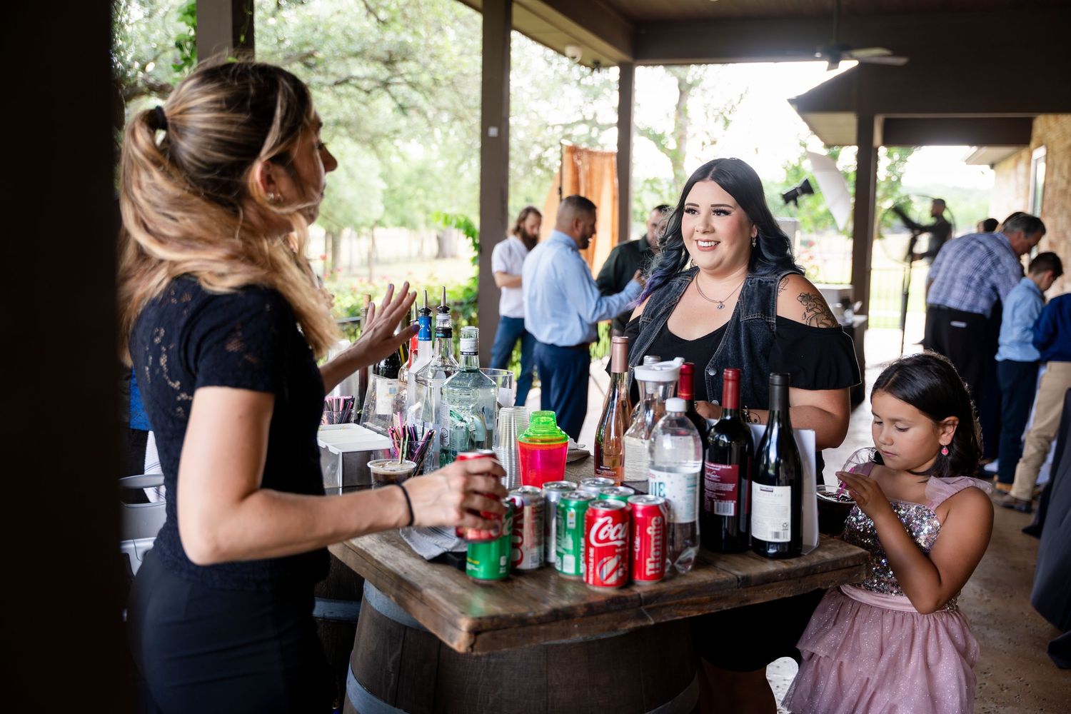 Party guests enjoy drinks and refreshments at a rustic covered outdoor venue with coke bottles and beverages on display.