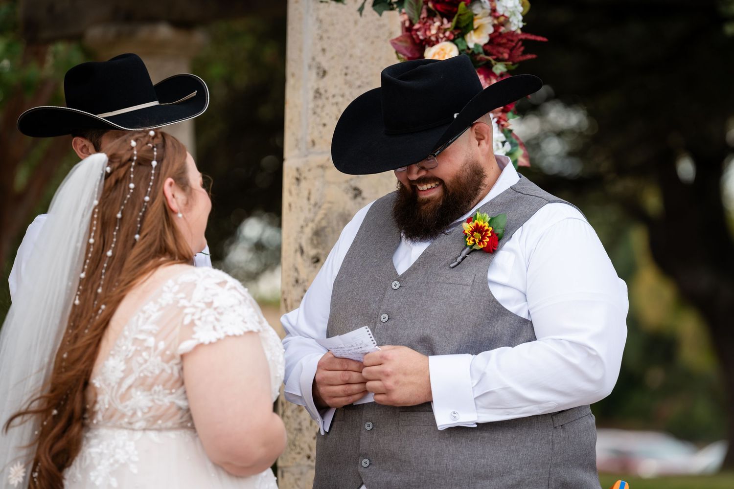 A wedding ceremony moment as the couple exchanges rings under a decorated arch while wearing western attire.