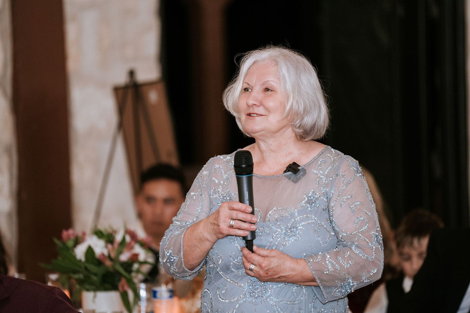An elegant senior woman in a sparkly gray dress gives a speech at a formal event.