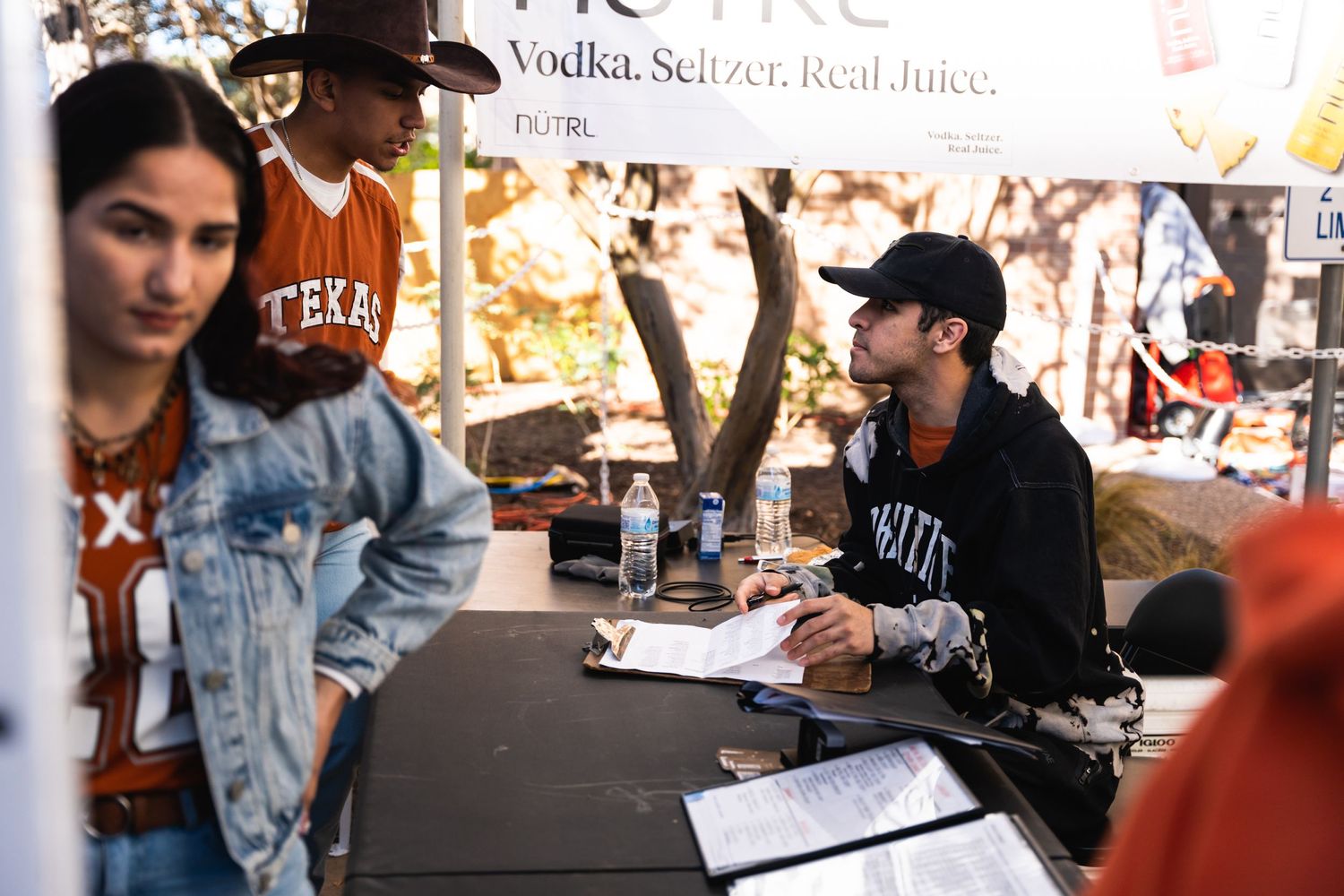 People at a merchandise signing table during an outdoor event with banners visible.