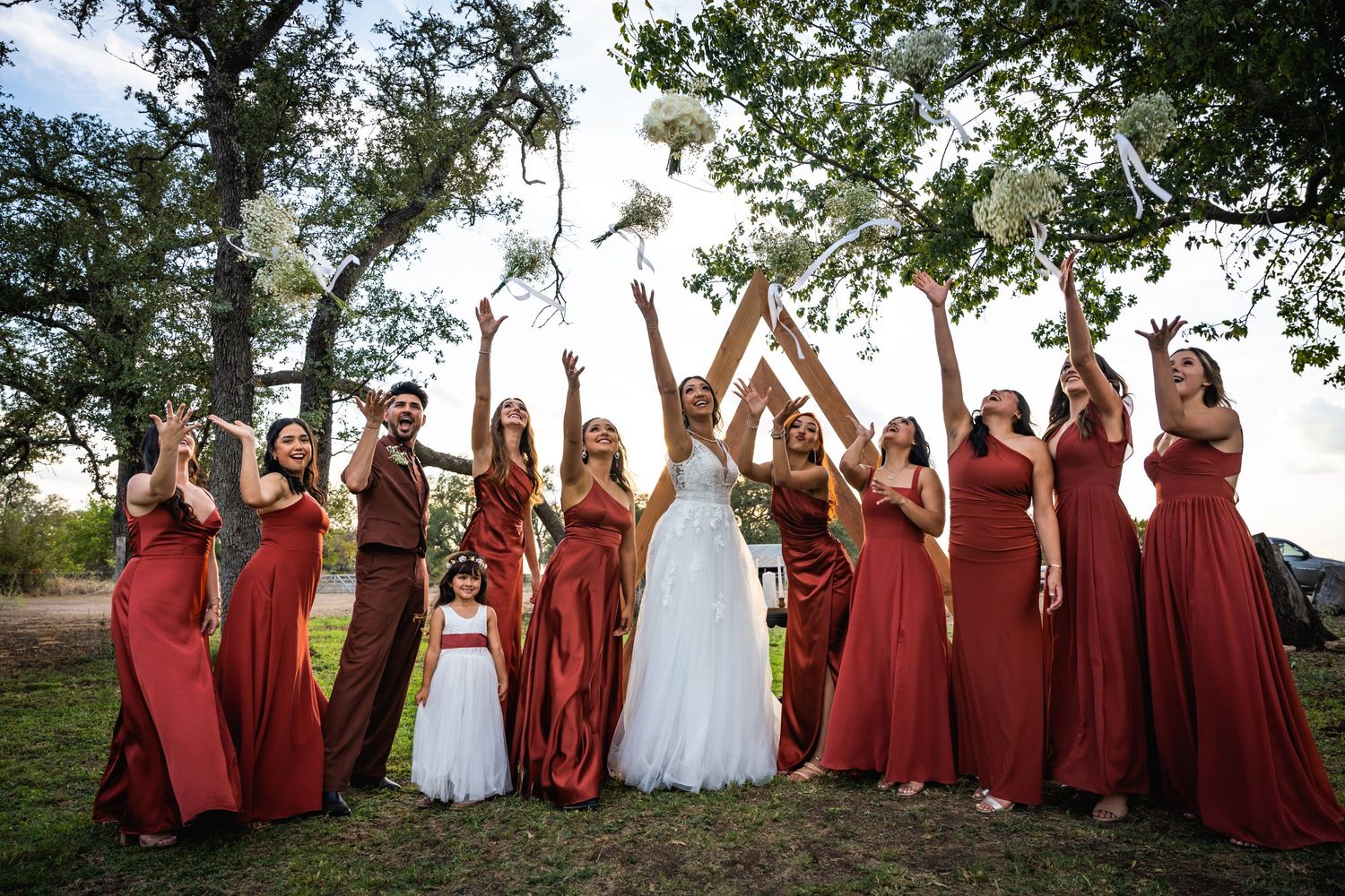 Bridesmaids in matching burgundy dresses celebrate with arms raised during outdoor wedding photo session.