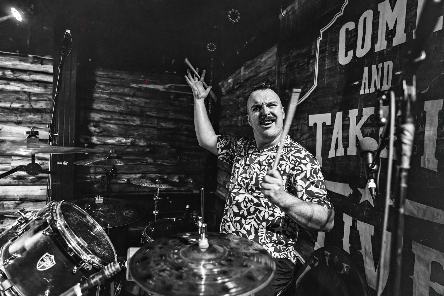 Black and white shot of performer enthusiastically playing drums on stage with rustic wooden backdrop.