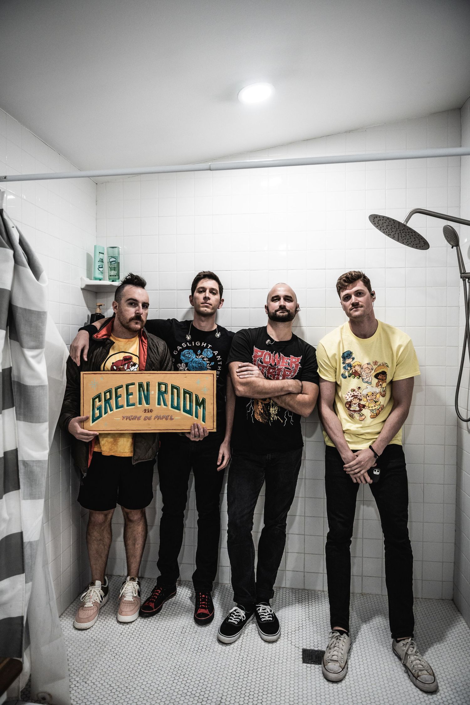 Band members posing in a bathroom holding a 'Green Room' sign in desaturated lighting.
