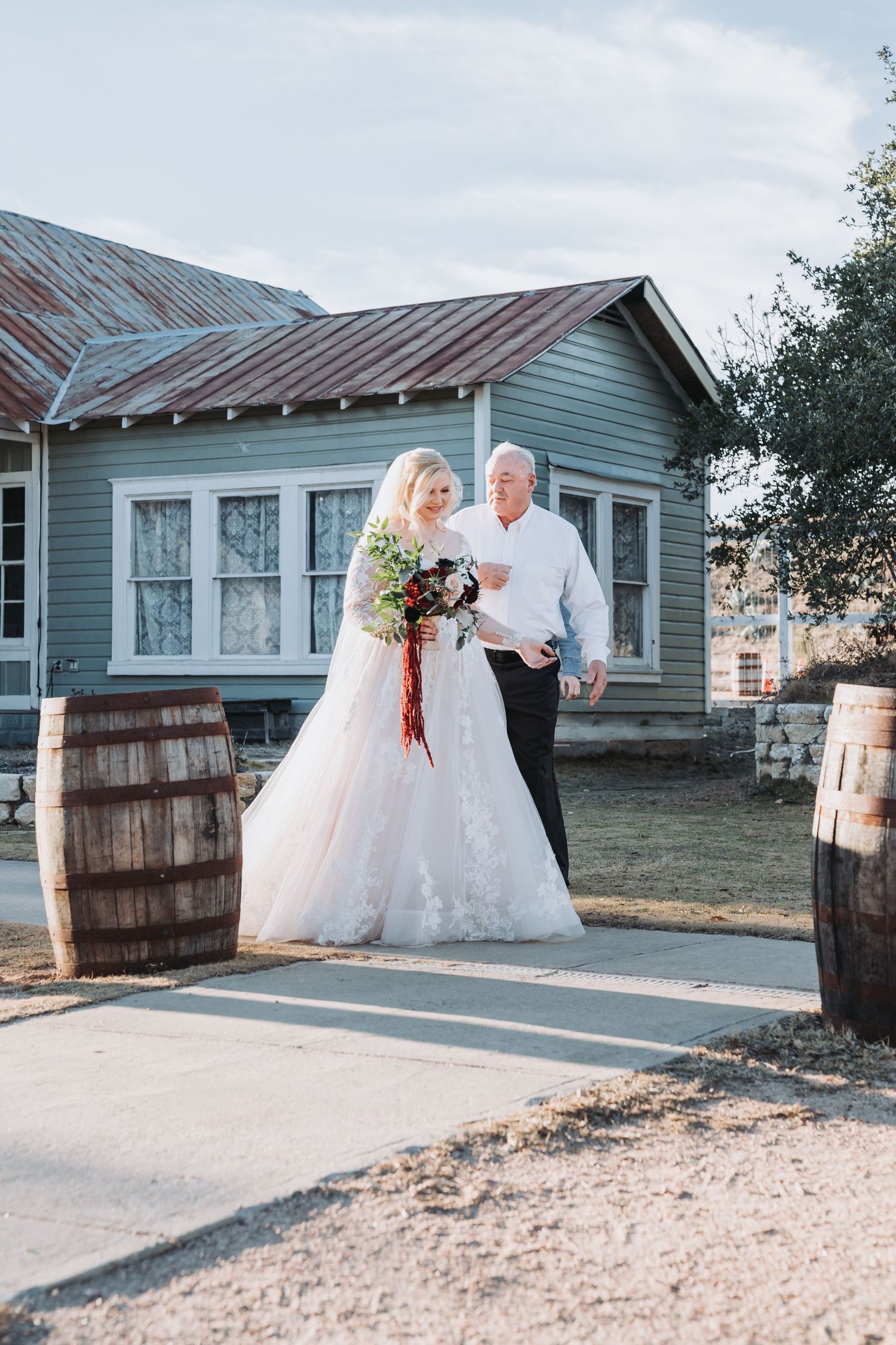 A bride in a white gown stands with companion by rustic wooden barrels.