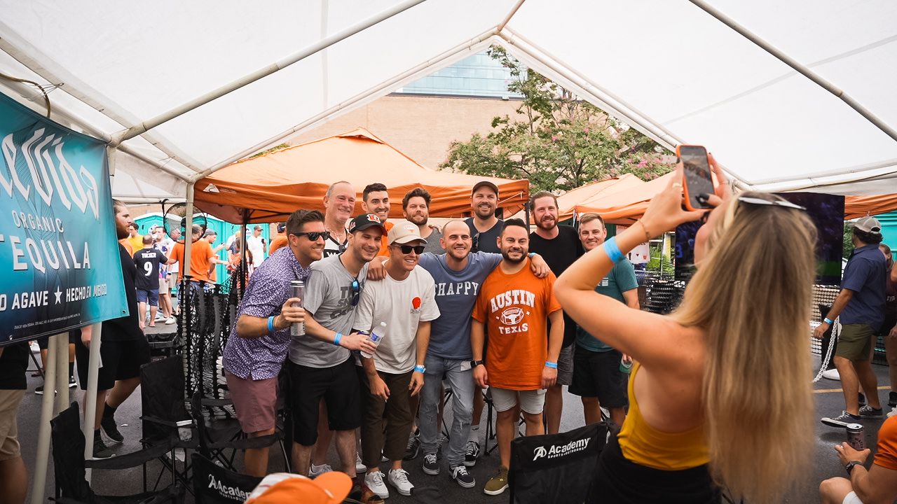 Energetic crowd gathers under white tent during outdoor event with orange-themed attire and decorations.