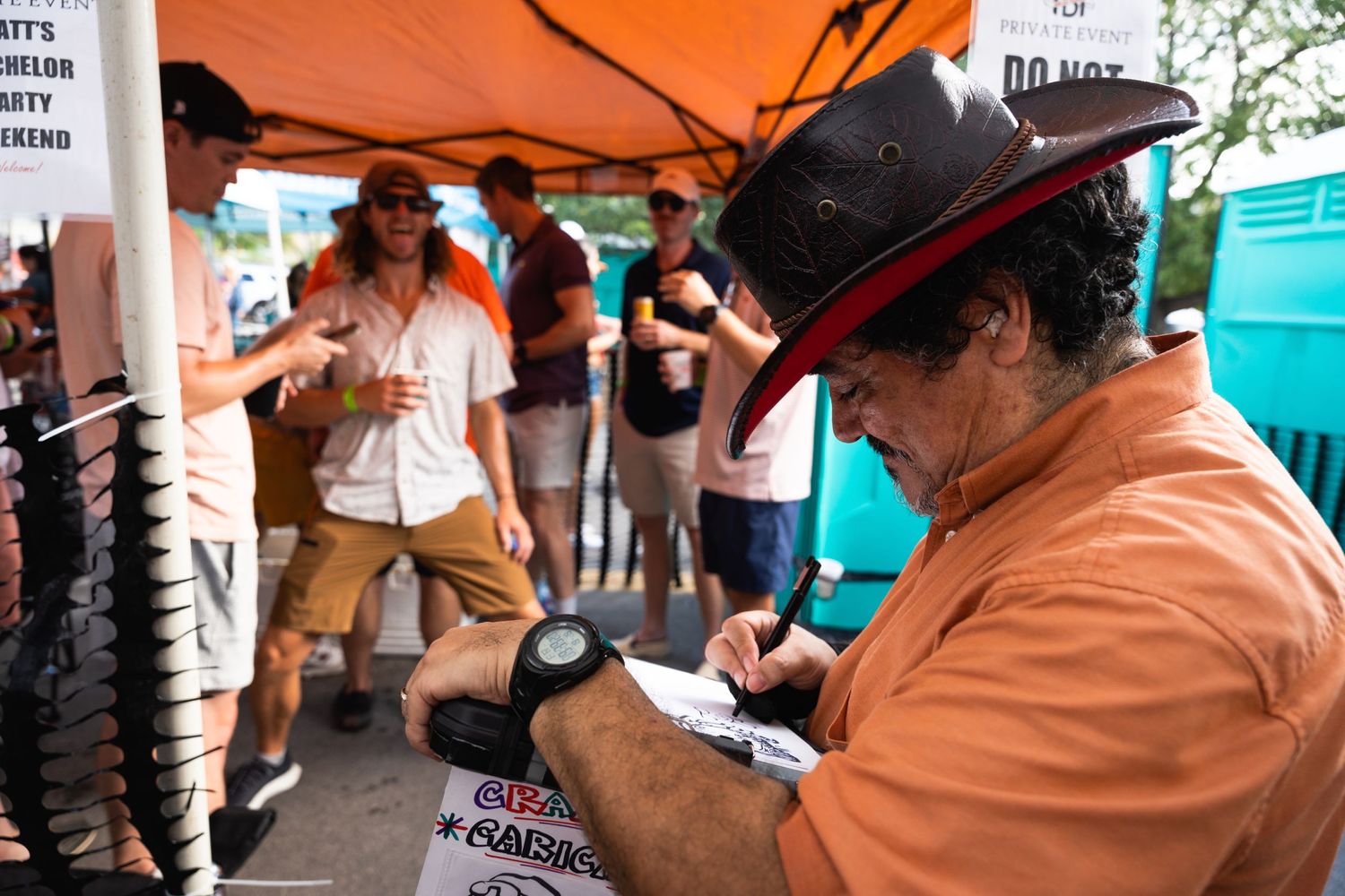 Person in cowboy hat signs or writes under orange tent at outdoor festival.