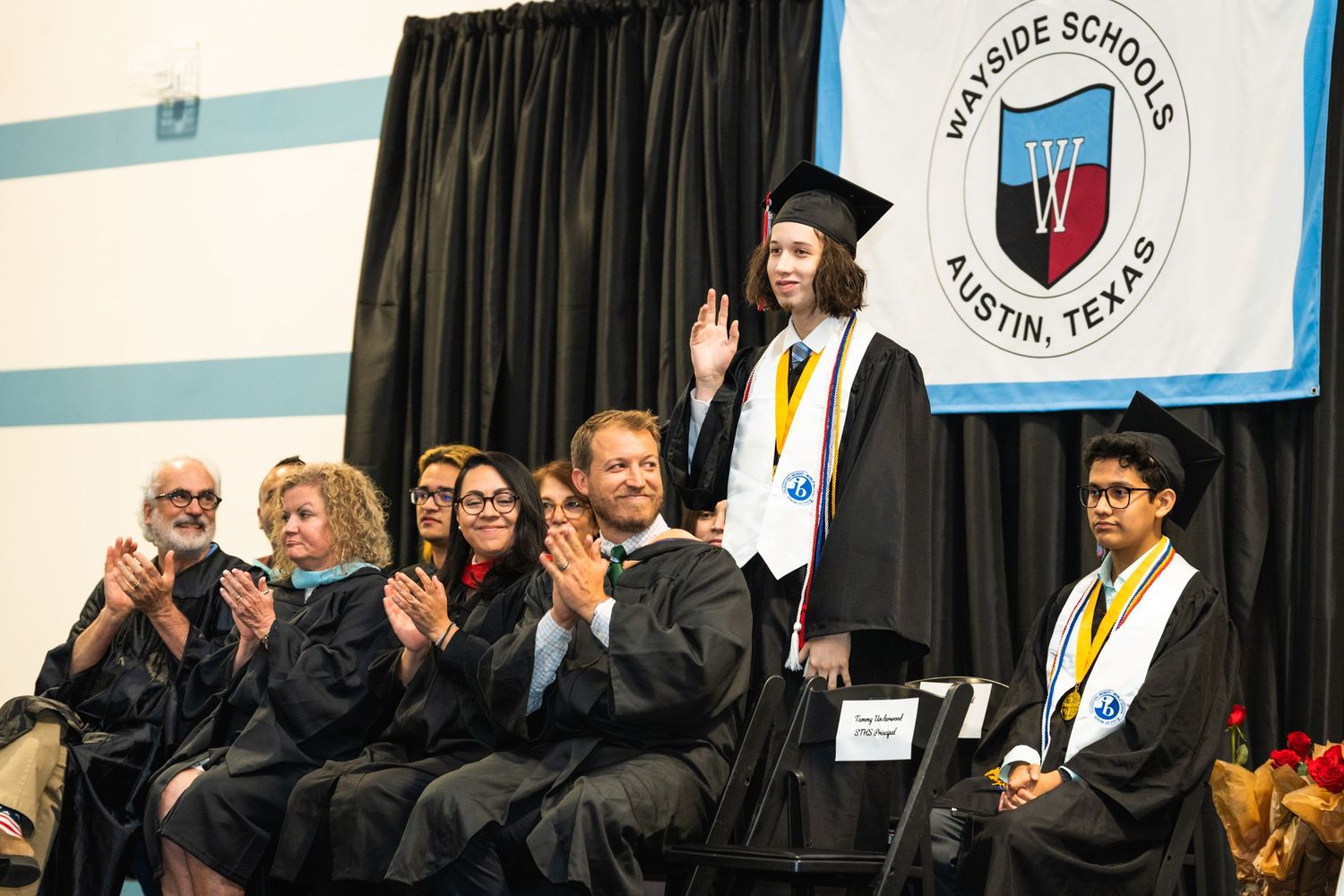 Graduation ceremony showing graduates in caps and gowns on stage with school banner.