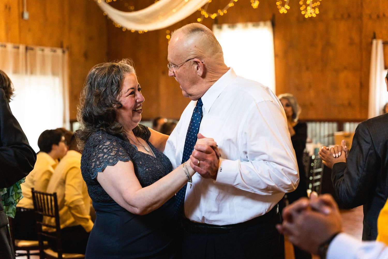 Couples slow dancing on wooden dance floor under string lights at wedding reception.