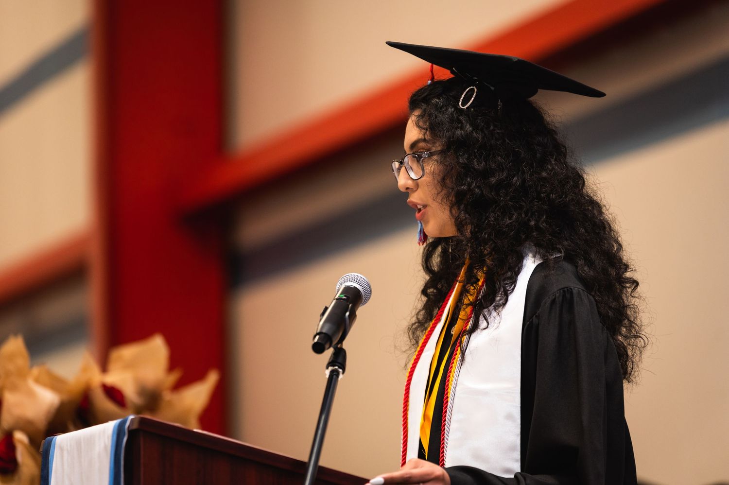 Graduate with curly hair giving commencement speech at podium.