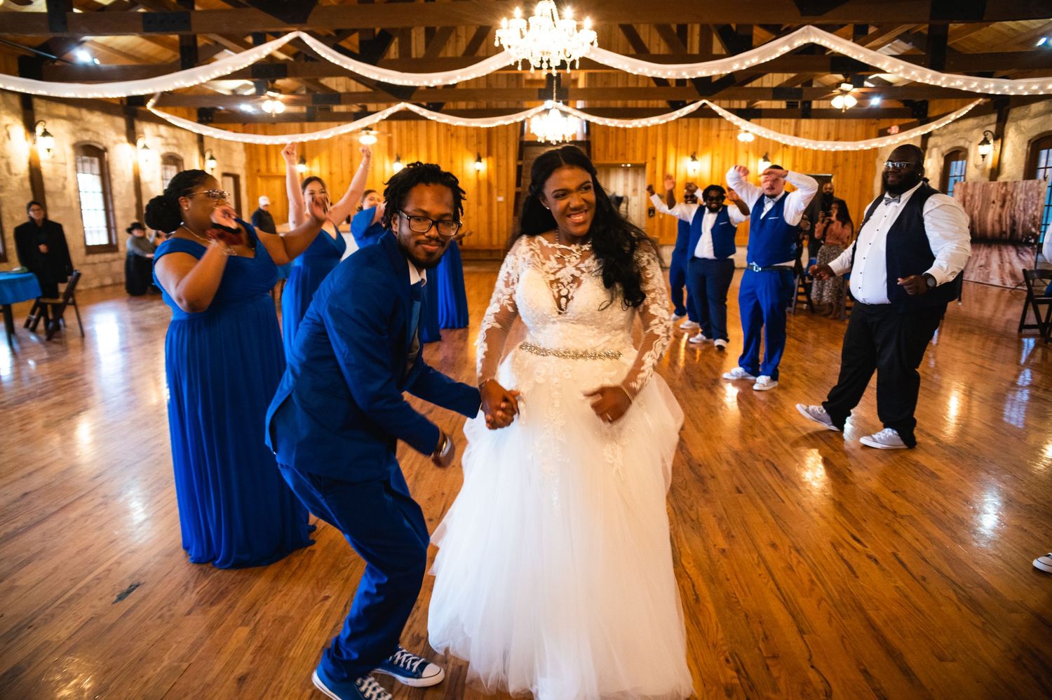 Wedding guests in blue dresses dance joyfully on a wooden dance floor decorated with elegant white draping and chandeliers.