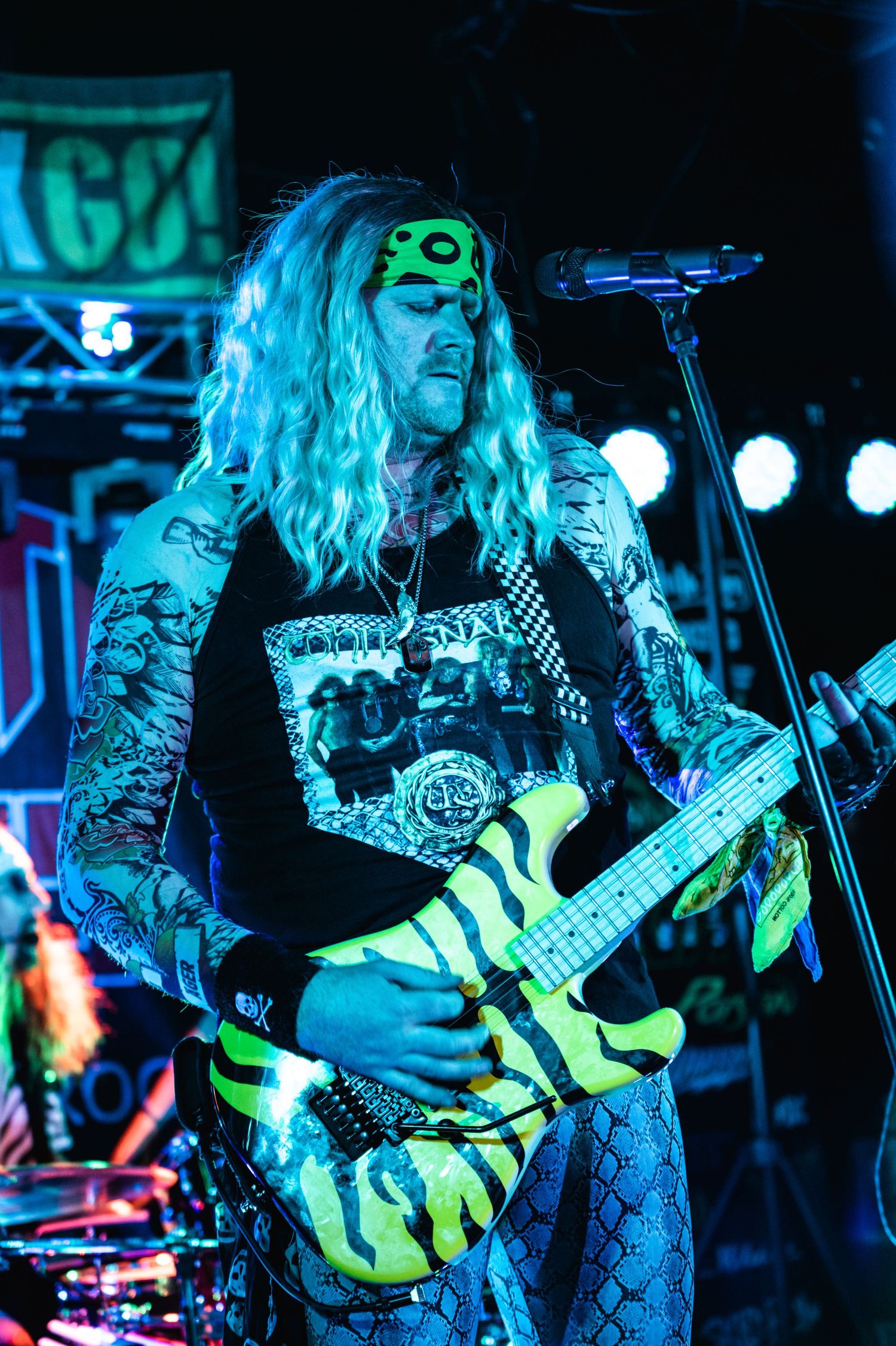Musician plays electric guitar with dramatic blue lighting and green face paint during performance.