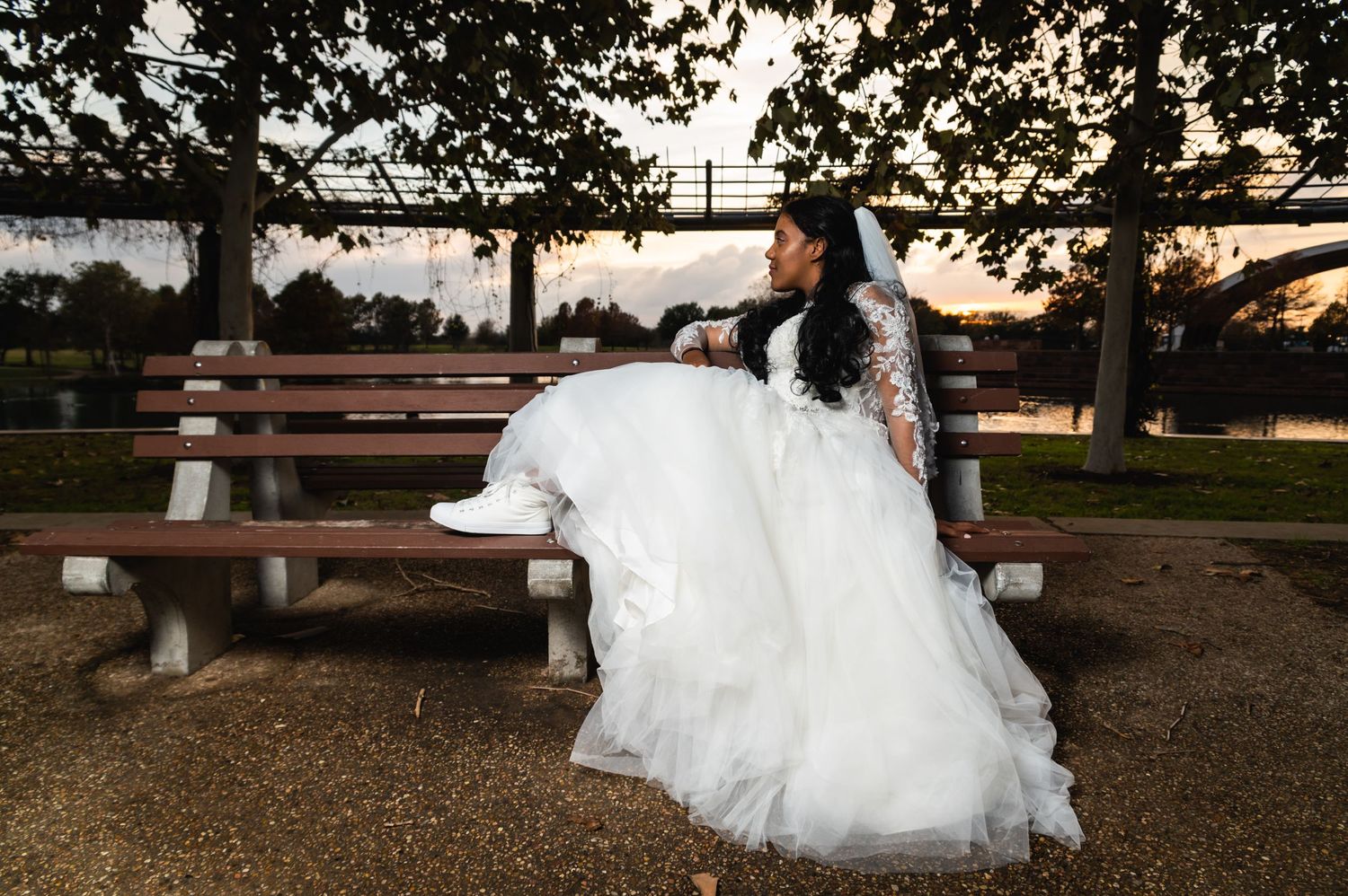 Bride in white gown sits gracefully on park bench at sunset.