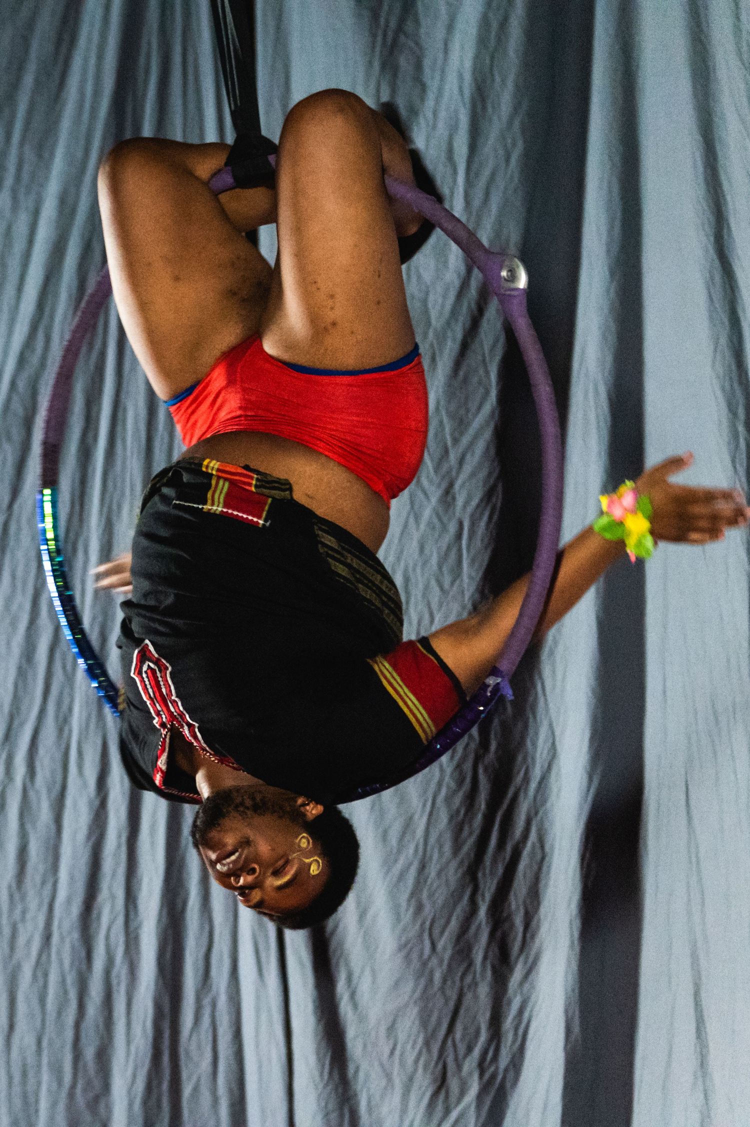 An aerial performer in red and black attire executes an inverted pose while suspended from purple aerial silks.