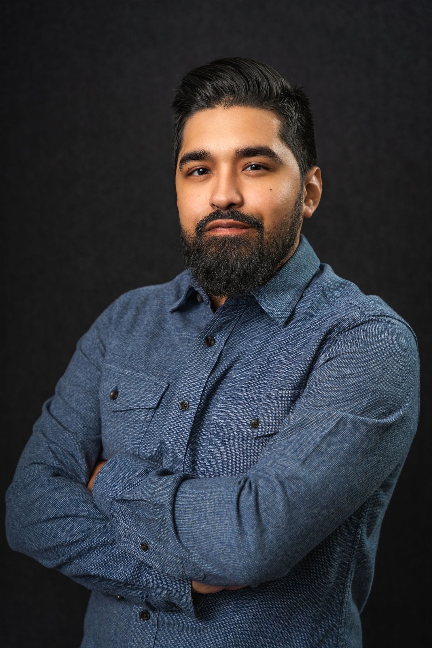 Professional portrait of person in blue denim shirt against dark background with confident pose and crossed arms.