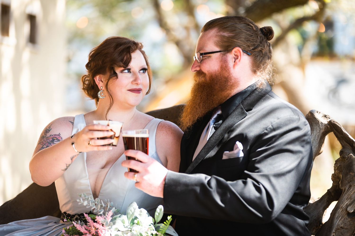 A couple shares a toast with beer glasses while celebrating their wedding day outdoors.