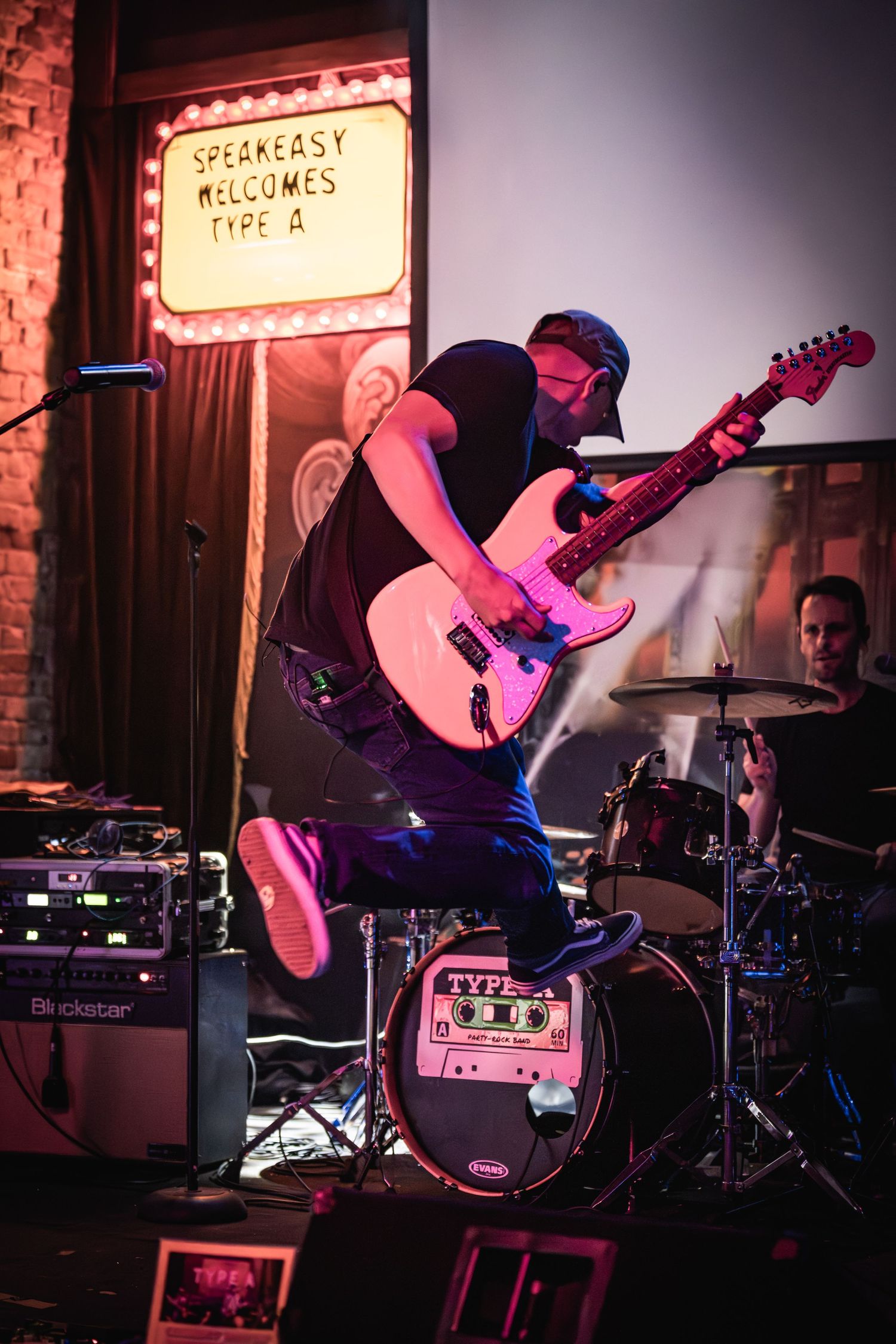 Energetic guitarist jumps while performing on stage with a pink telecaster under dramatic lighting.