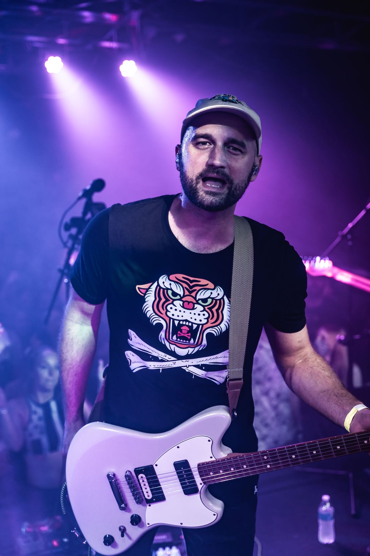 Musician performs on stage wearing a tiger graphic t-shirt while playing a white electric guitar.