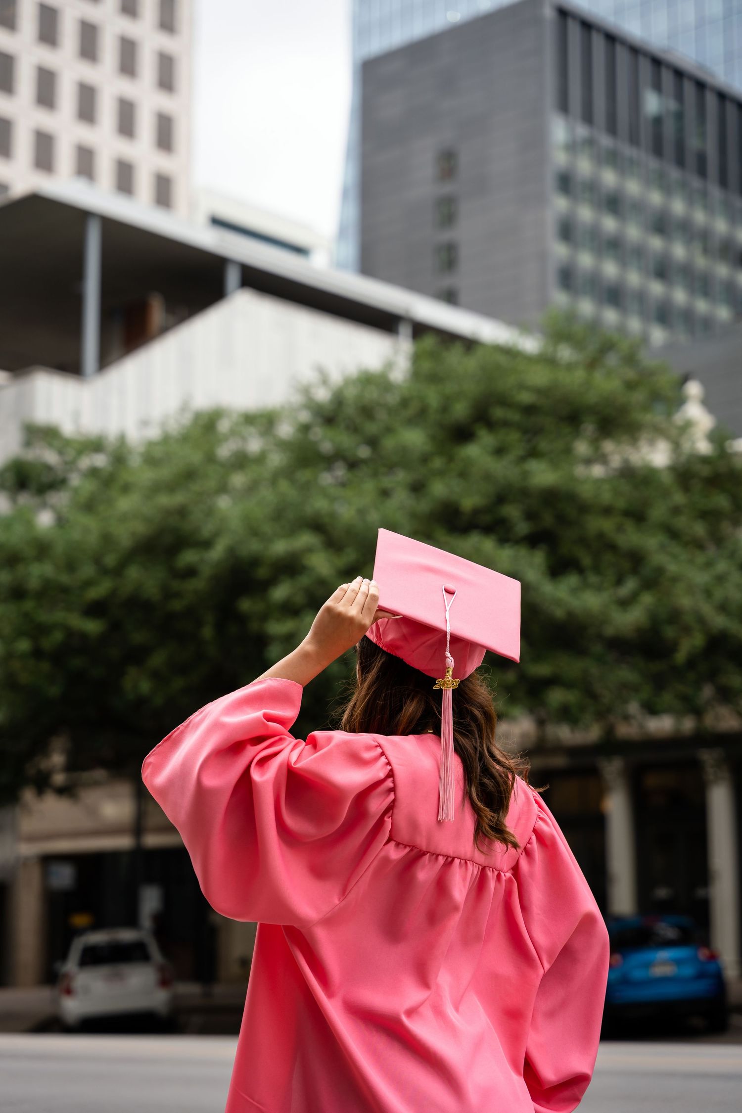 Graduate in pink graduation gown adjusts cap against urban backdrop of tall buildings.