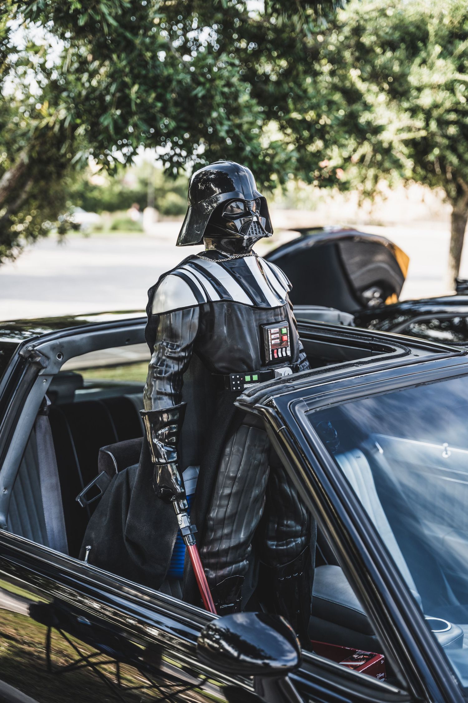 Darth Vader costume emerging from black car sunroof with lightsaber.